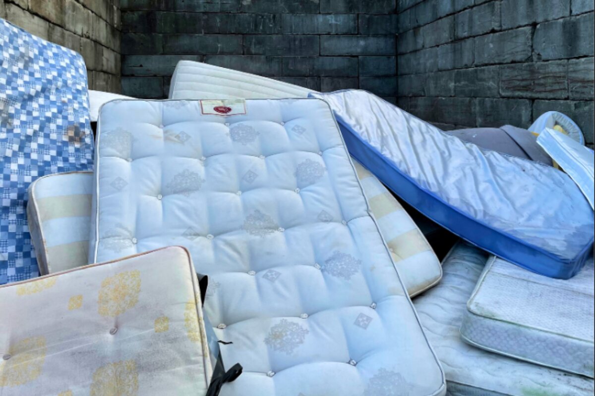 how-get-rid-king-size-mattress-what-to-do-with-king-size-bed-frame stack of used mattresses piled in a dumpster or disposal area against a brick wall.