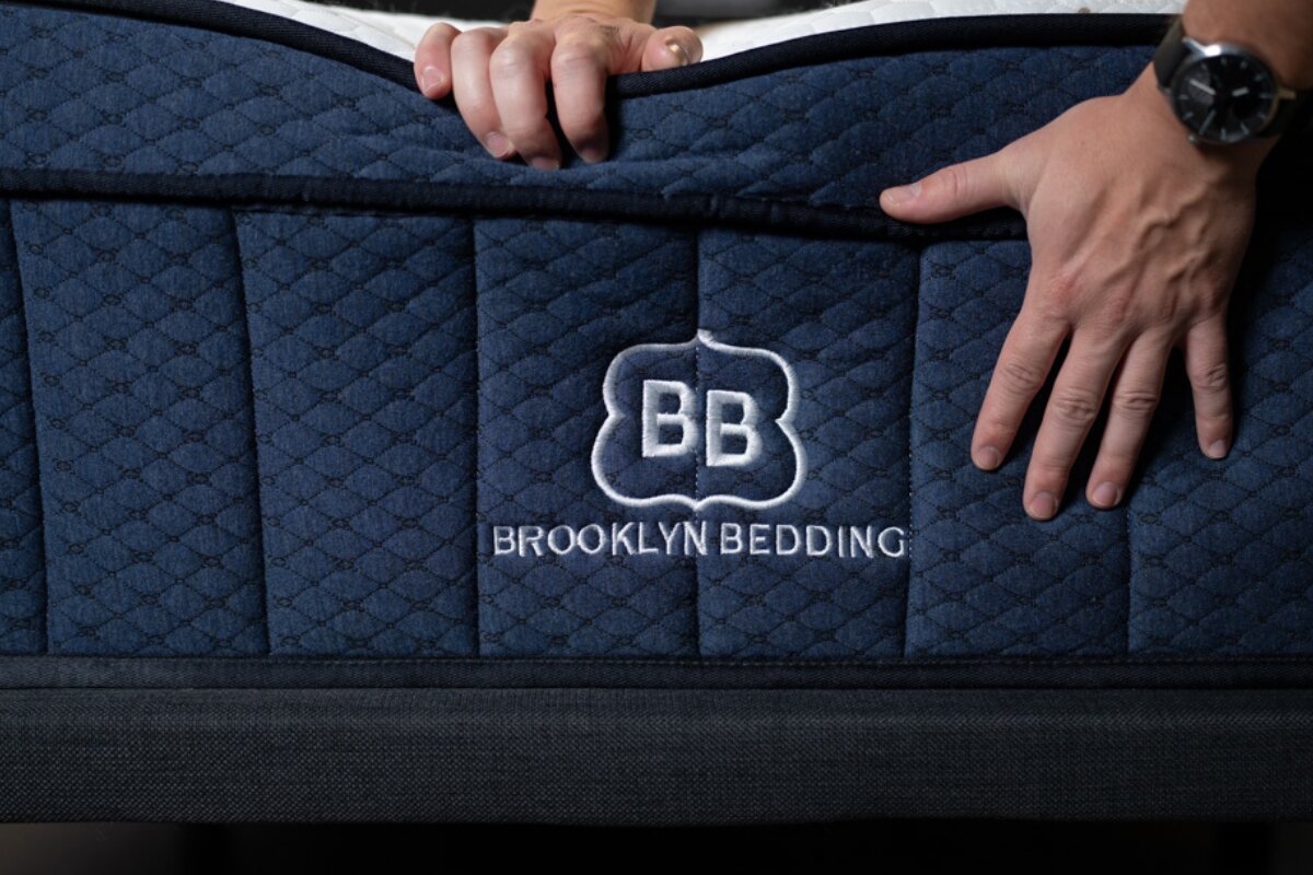 Brooklyn Bedding mattress close-up showing quilted side panel and brand logo being pressed by hands