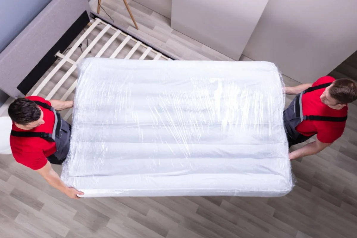 Two movers carrying a wrapped full size mattress through a bedroom during transport