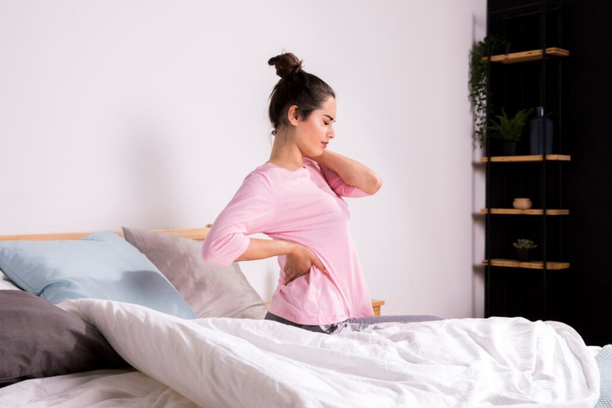 Woman sitting on a bed with lower back pain showing how mattress firmness can affect spinal support and sleep comfort