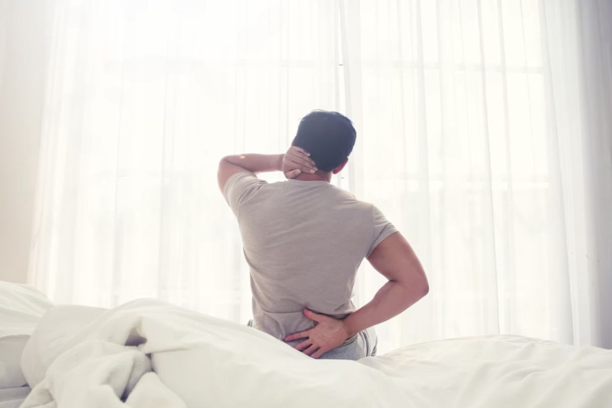 Man sitting on a bed with lower back and neck pain showing how mattress firmness can affect spinal support and comfort