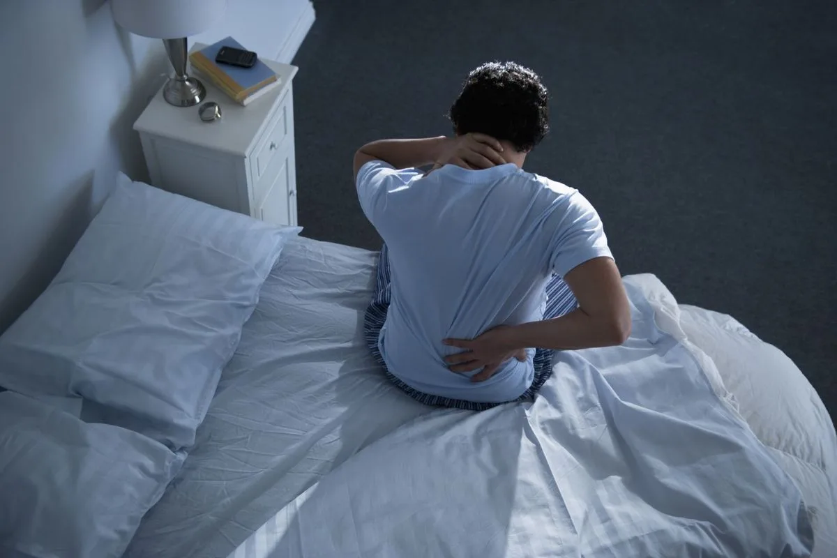 Person sitting on the edge of a bed while holding their lower back, illustrating how the wrong mattress can contribute to back pain and morning discomfort