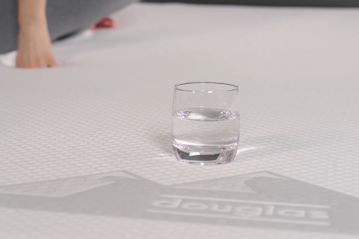 A glass of water on a memory foam bed with a person pushing on the mattress in the background.