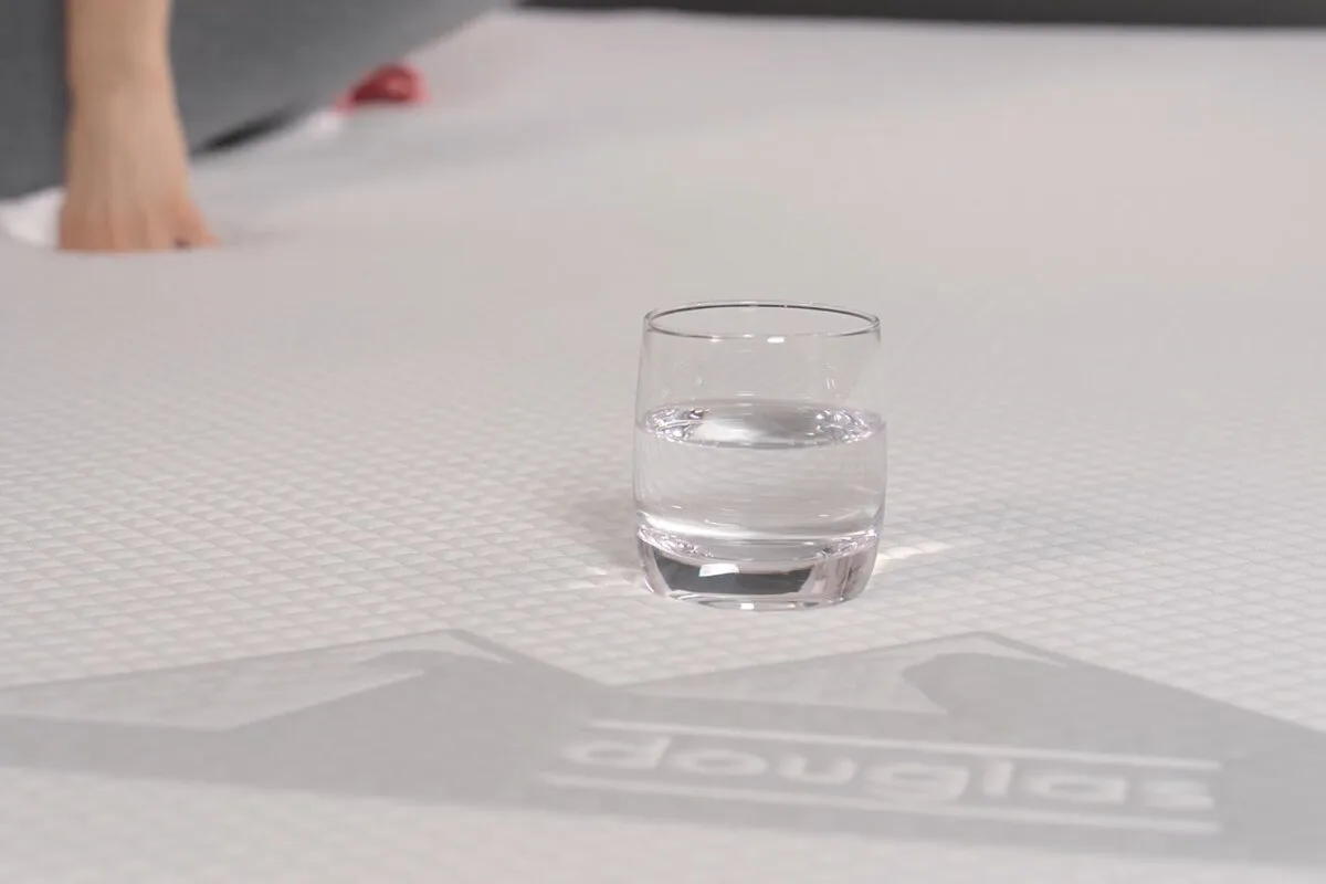 A glass of water on a memory foam bed with a person pushing on the mattress in the background.
