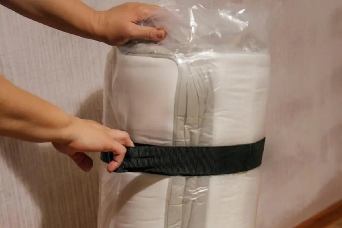 Person securing a compressed hybrid mattress with straps to keep it tightly rolled before transport or storage.