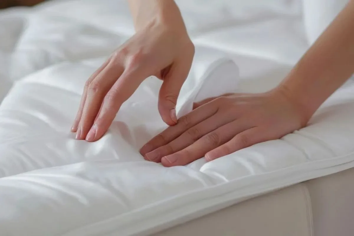 Hands placing a quilted mattress pad on a bed, illustrating the use of breathable mattress toppers and protectors to help keep a memory foam mattress cooler