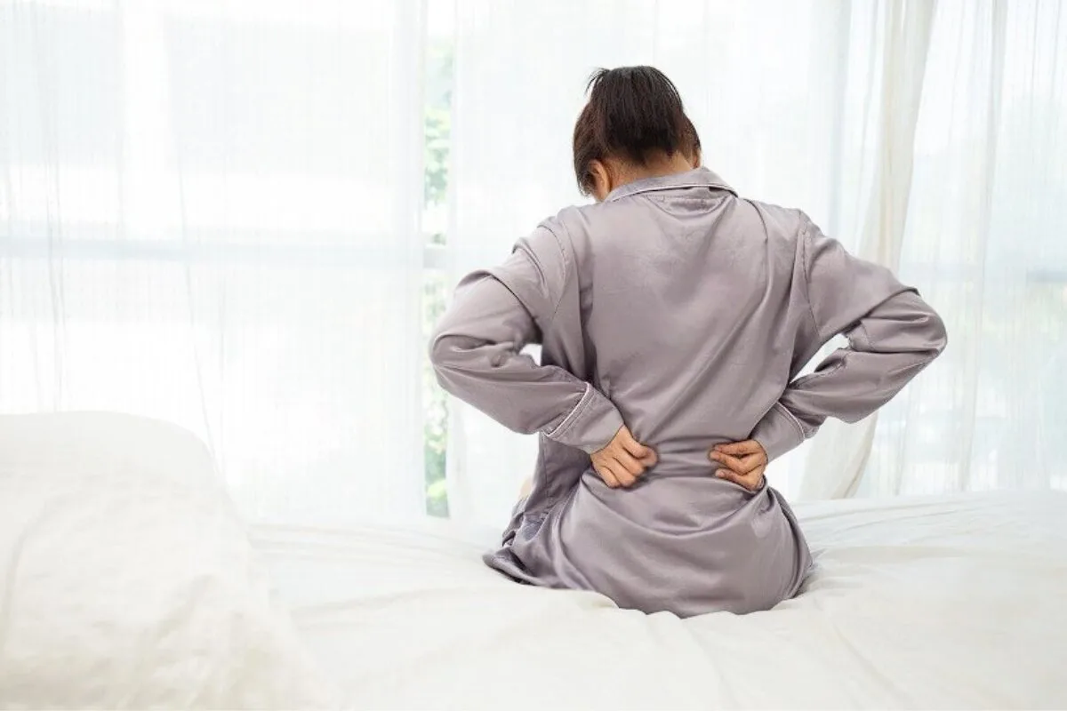 A woman holding on to her back in pain while sitting at the edge of the bed.