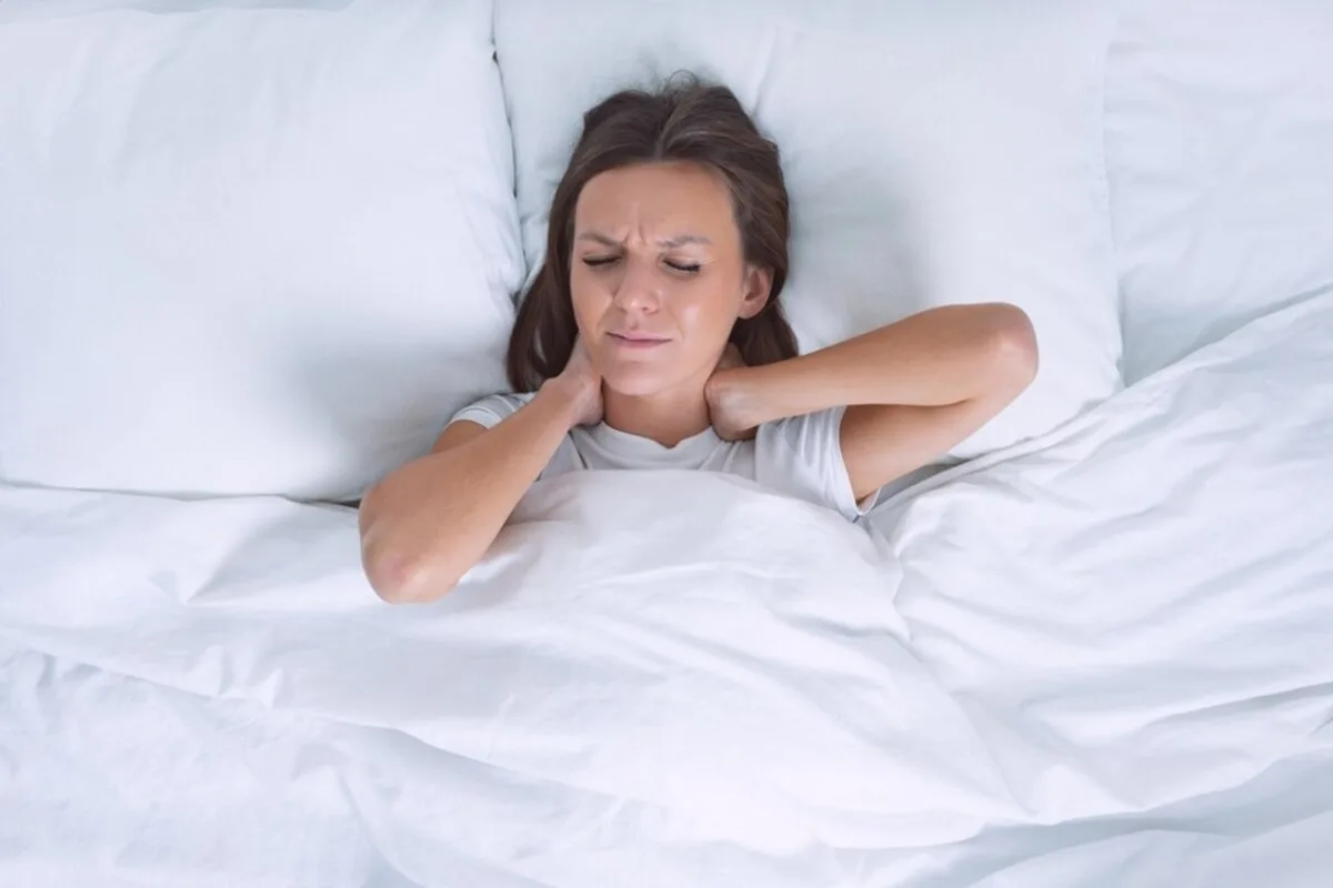 What Mattress Firmness Is Best for Stomach Sleepers? A woman with neck strain lying down on her back in a bed.