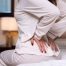 Person sitting on a bed holding their lower back, illustrating back pain that can result from stomach sleeping and poor spinal alignment