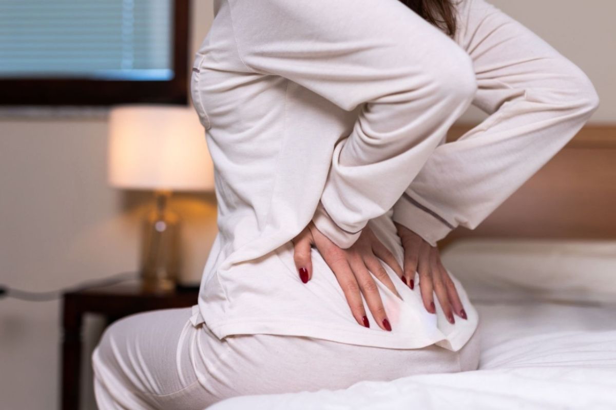 Person sitting on a bed holding their lower back, illustrating back pain that can result from stomach sleeping and poor spinal alignment